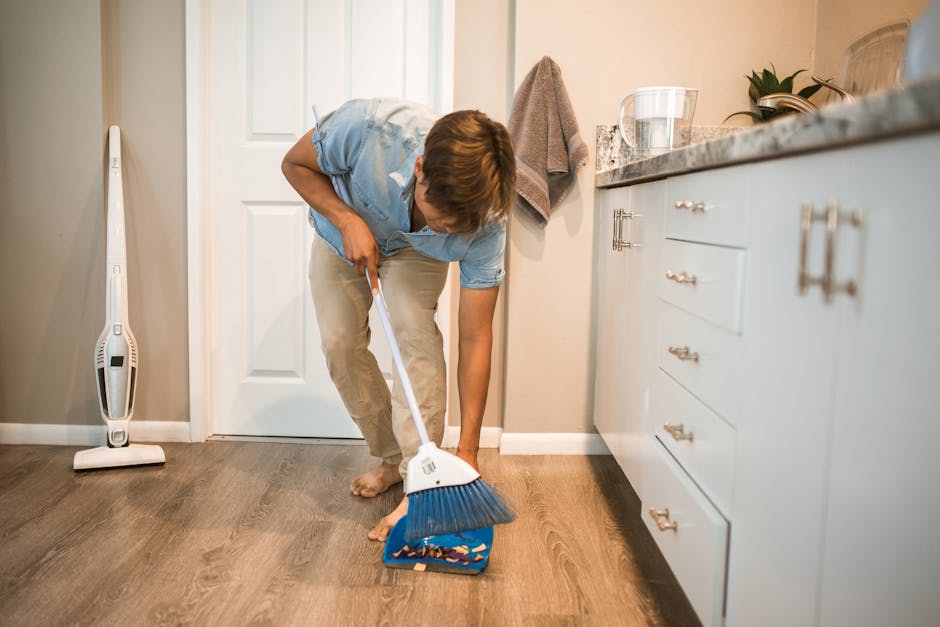 Man cleaning kitchen with a broom and dustpan on a wooden floor.
